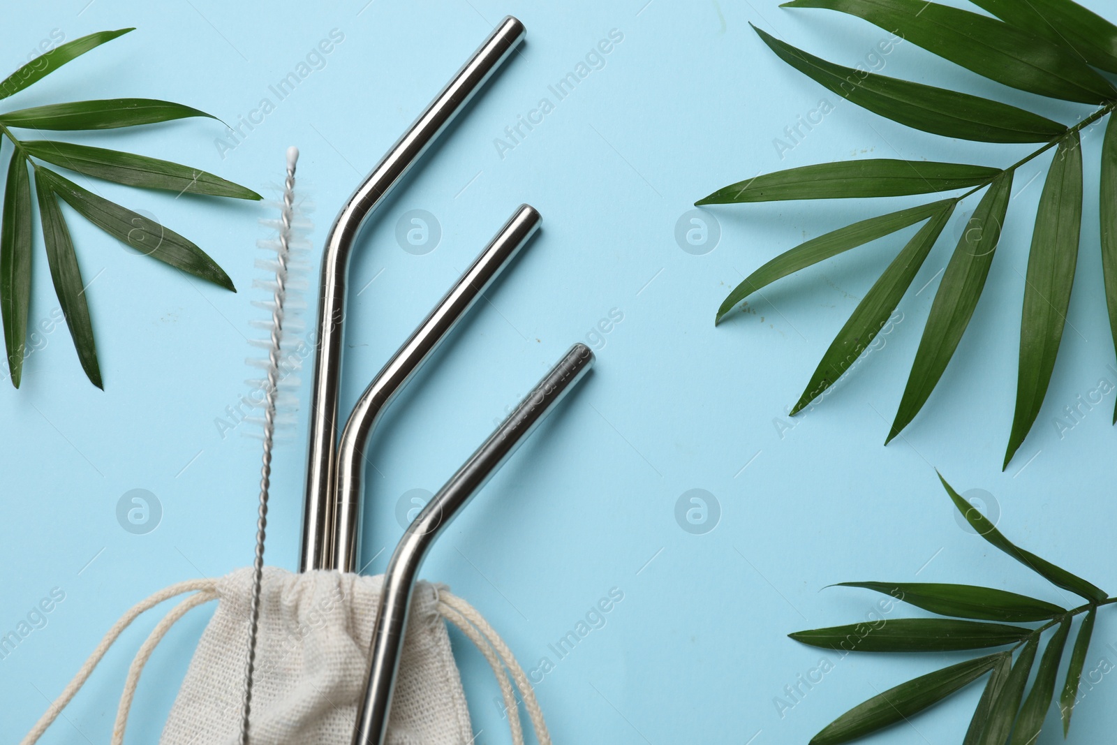 Photo of Metal drinking straws, cleaning brush, bag and palm leaves on light blue background, flat lay