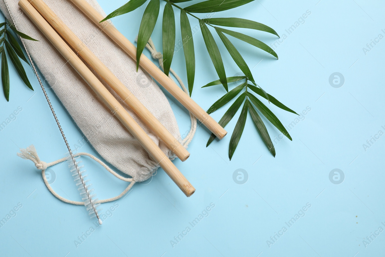 Photo of Bamboo drinking straws, cleaning brush, bag and palm leaves on light blue background, flat lay. Space for text