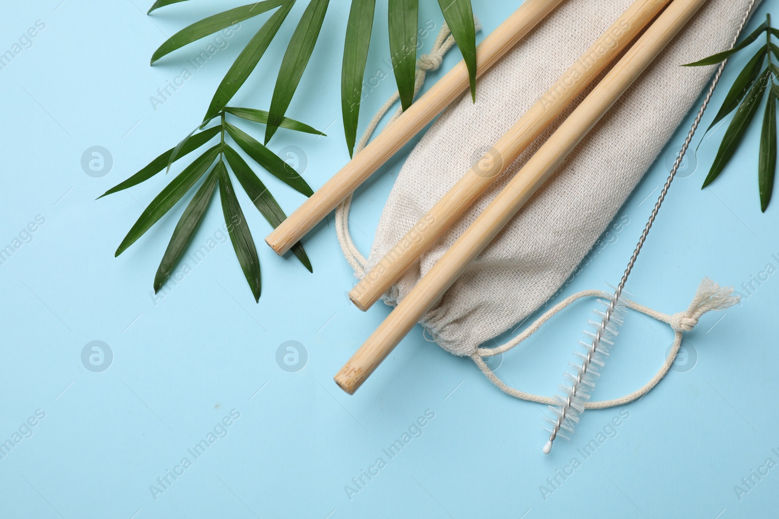 Photo of Bamboo drinking straws, cleaning brush, bag and palm leaves on light blue background, flat lay. Space for text