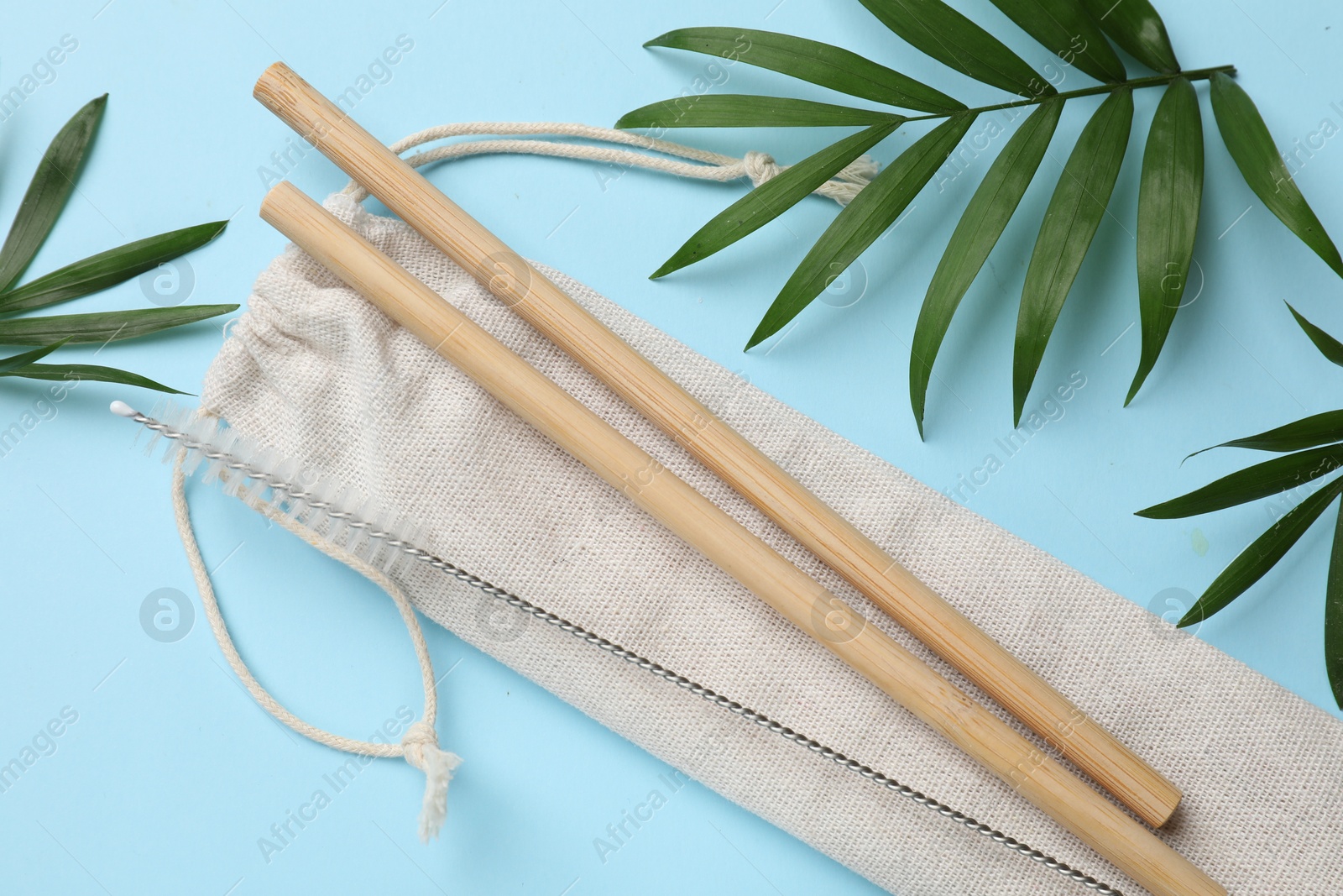 Photo of Bamboo drinking straws, cleaning brush, bag and palm leaves on light blue background, flat lay