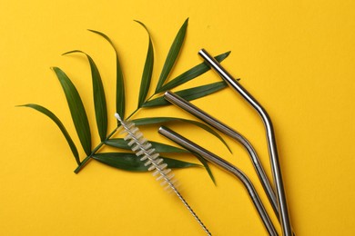 Photo of Metal drinking straws, cleaning brush and palm leaf on yellow background, flat lay