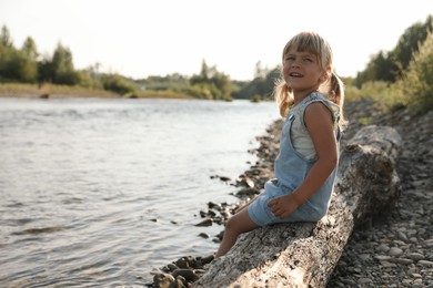 Cute little girl sitting on tree trunk near river. Child enjoying beautiful nature