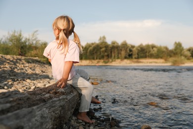 Photo of Cute little girl sitting on tree trunk near river. Child enjoying beautiful nature