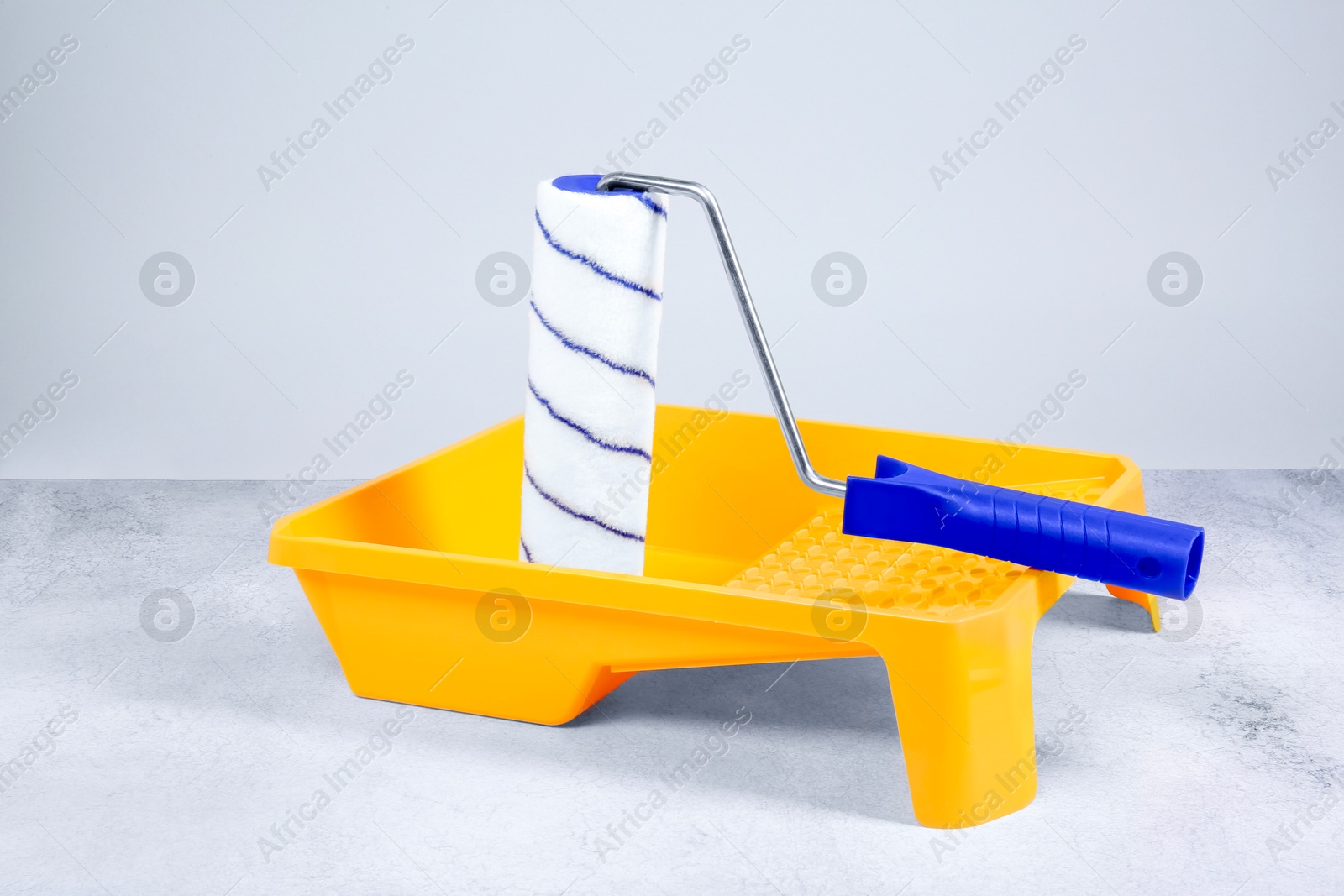 Photo of Paint roller and yellow tray on textured table against light background. Building material