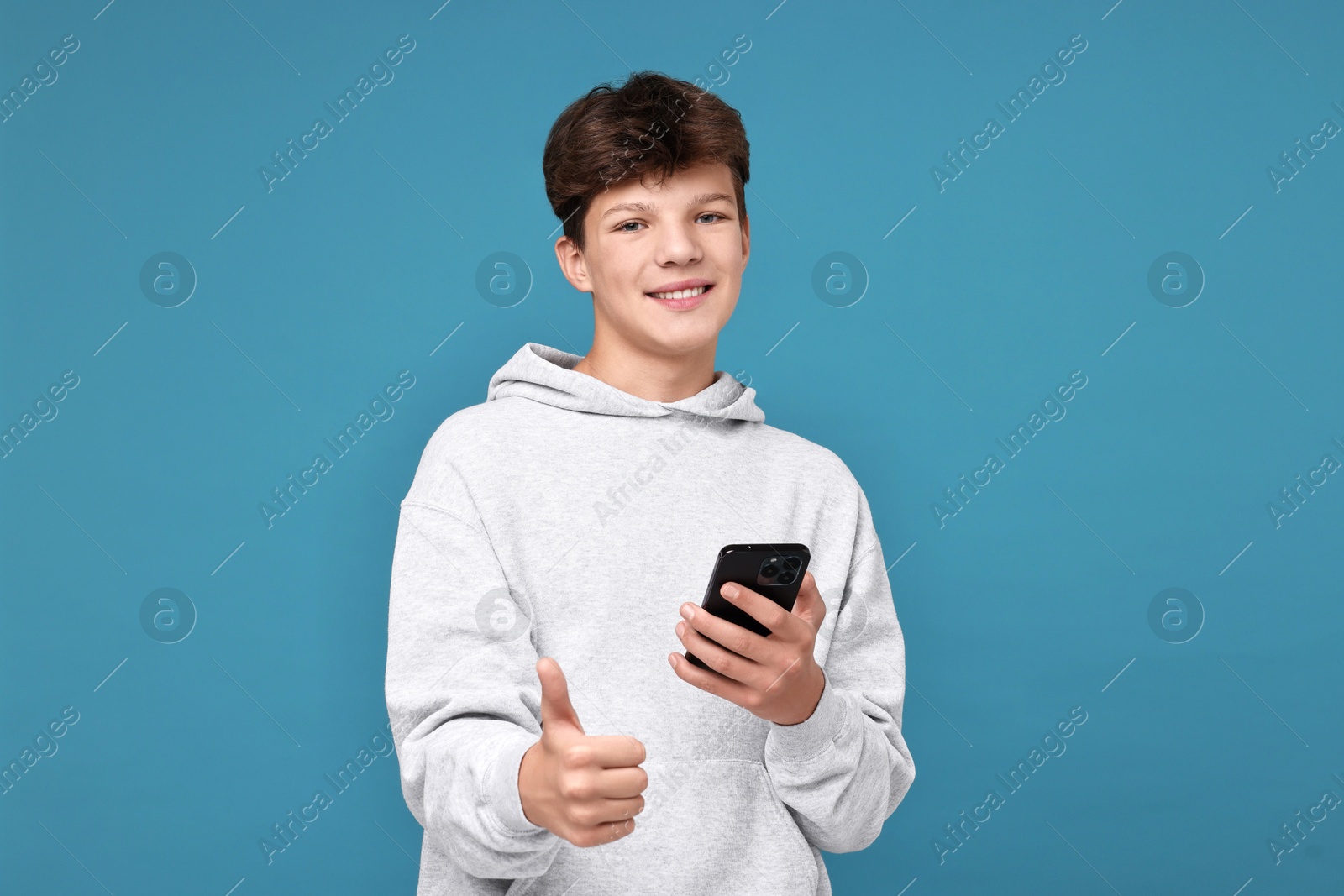 Photo of Teenage boy with smartphone showing thumbs up on light blue background