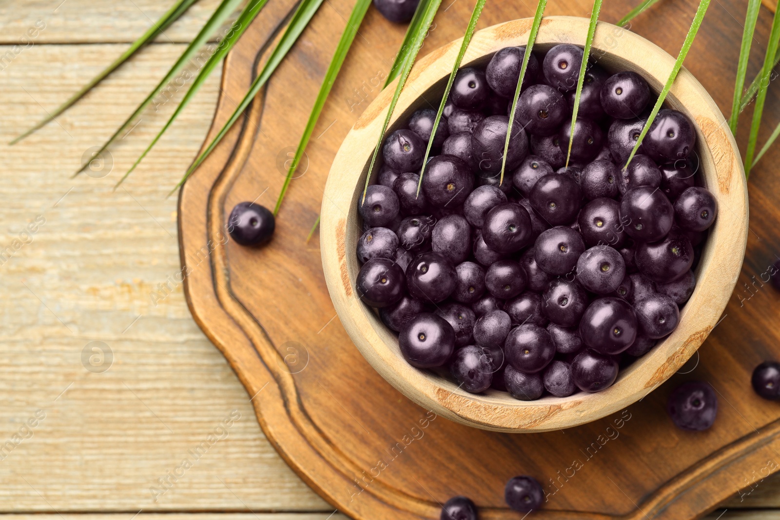 Photo of Ripe acai berries in bowl and palm leaves on wooden table, flat lay