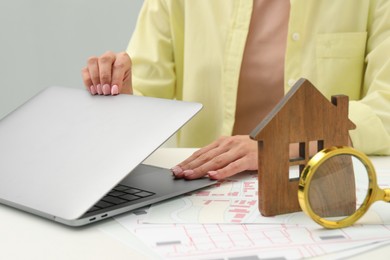 Photo of House hunting. Woman with laptop, papers, magnifying glass and house figure at white table, closeup