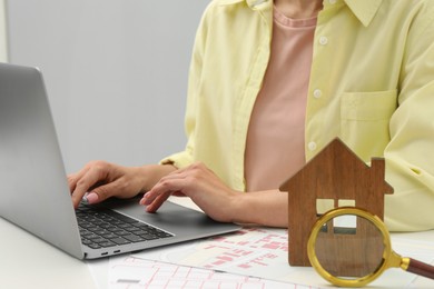 Photo of House hunting. Woman with laptop, papers, magnifying glass and house figure at white table, closeup