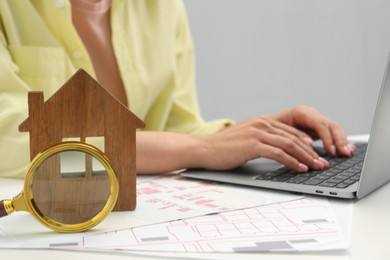 Photo of House hunting. Woman with laptop, papers, magnifying glass and house figure at white table, closeup