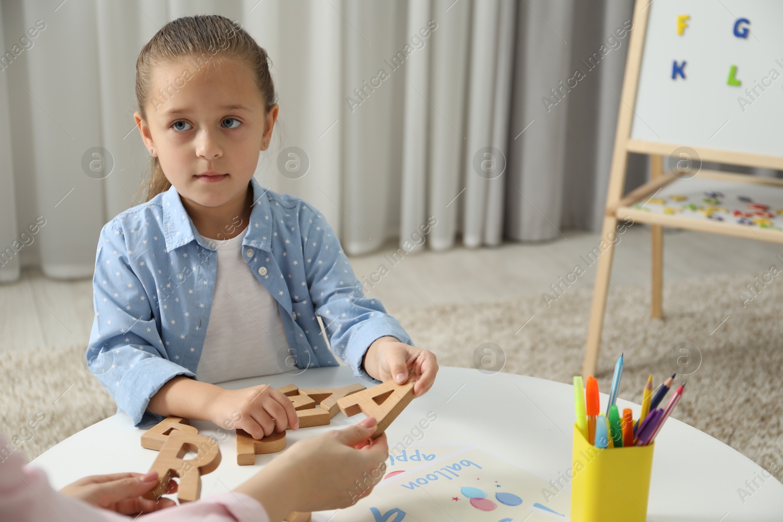 Photo of Speech therapist teaching little girl alphabet at white table indoors, closeup