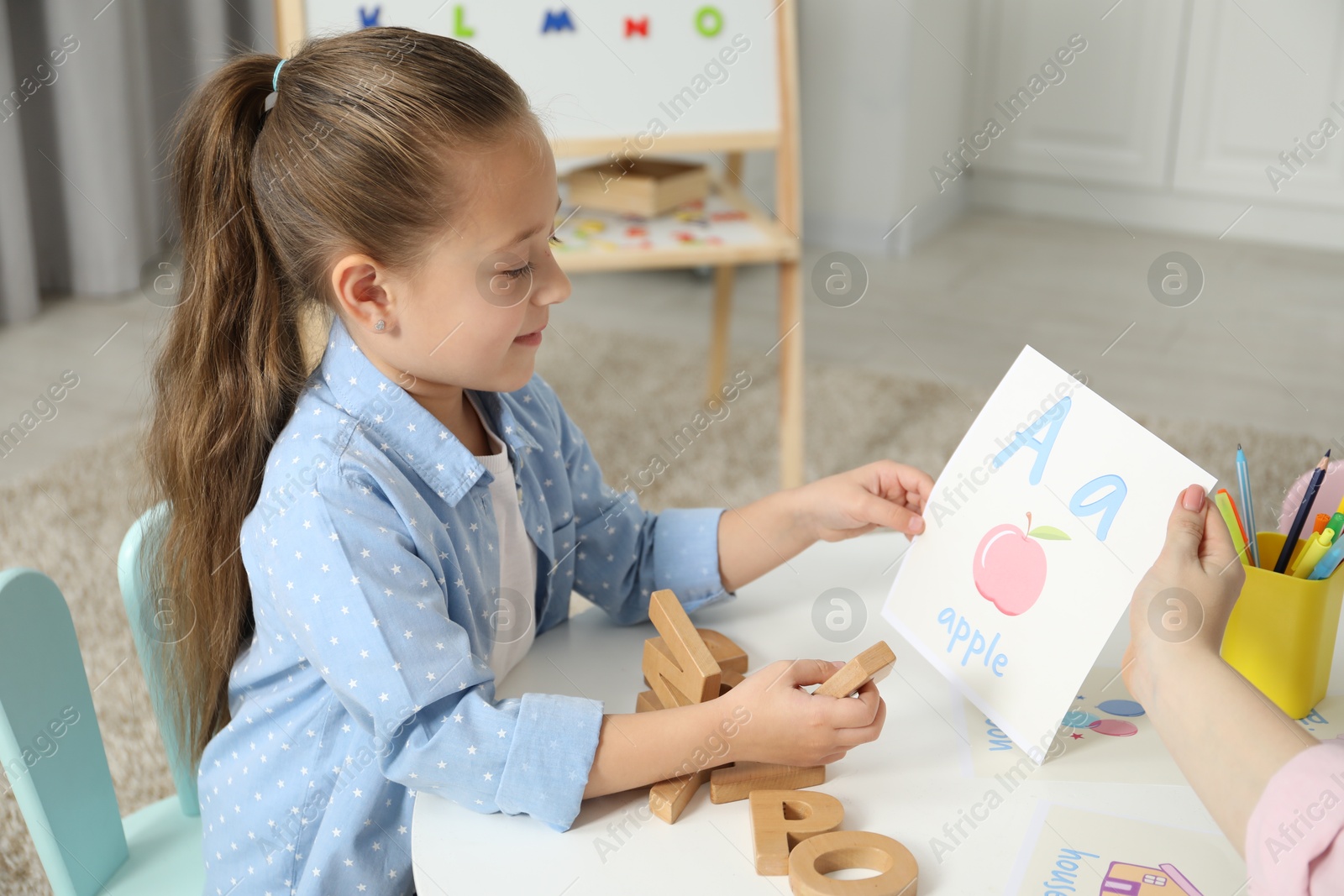 Photo of Speech therapist teaching little girl alphabet at white table indoors, closeup