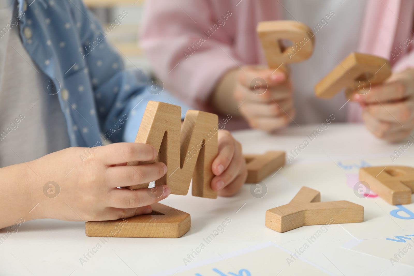 Photo of Speech therapist teaching little girl alphabet with wooden letters at white table indoors, closeup