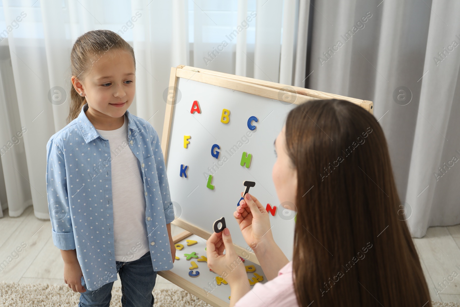 Photo of Speech therapist teaching little girl alphabet with magnetic letters indoors