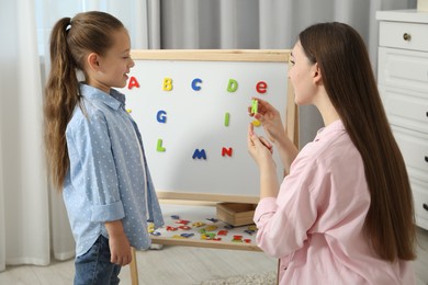 Photo of Speech therapist teaching little girl alphabet with magnetic letters indoors