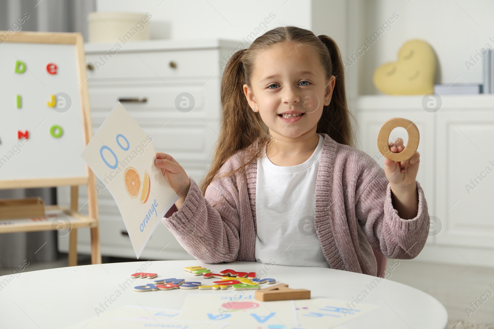 Photo of Little girl learning alphabet with different letters and picture at white table indoors