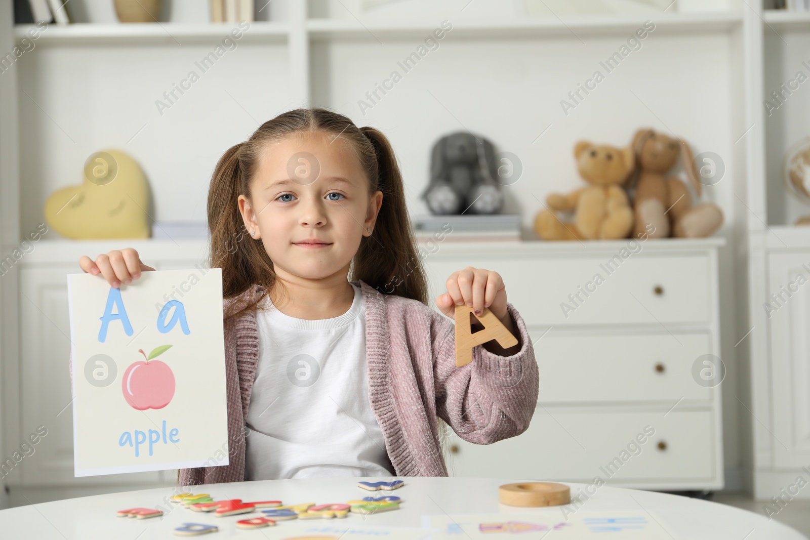 Photo of Little girl learning alphabet with different letters and picture at white table indoors