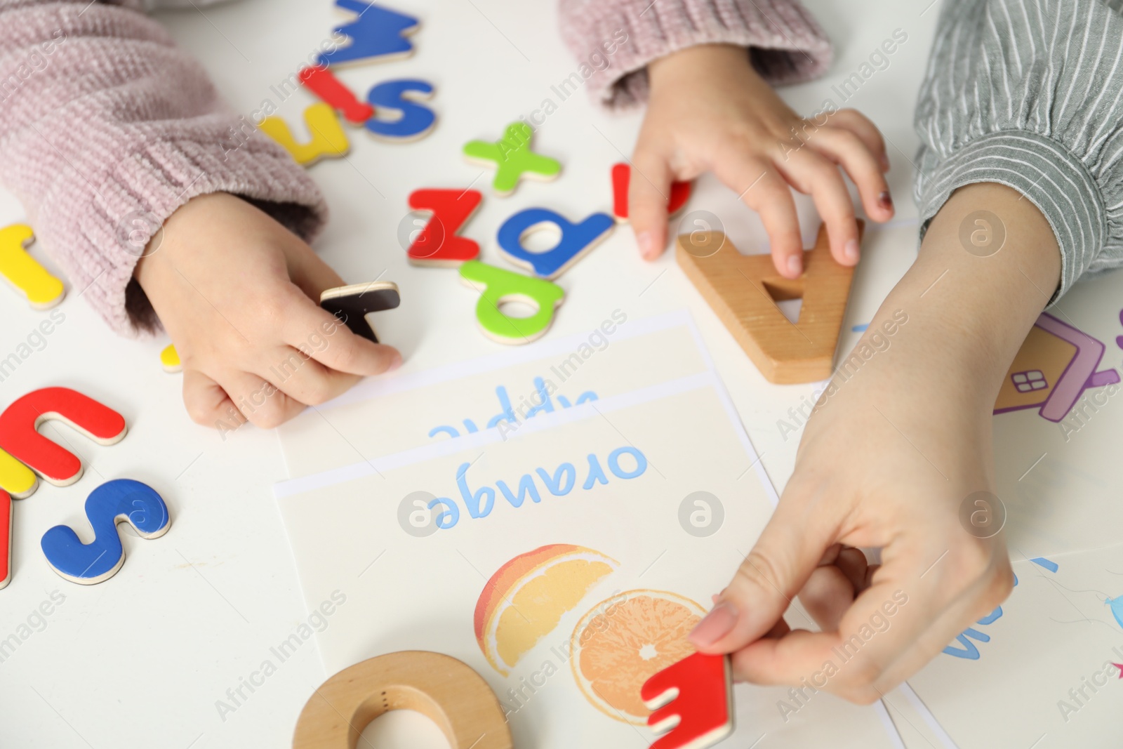 Photo of Speech therapist teaching little girl alphabet with different letters at white table indoors, closeup