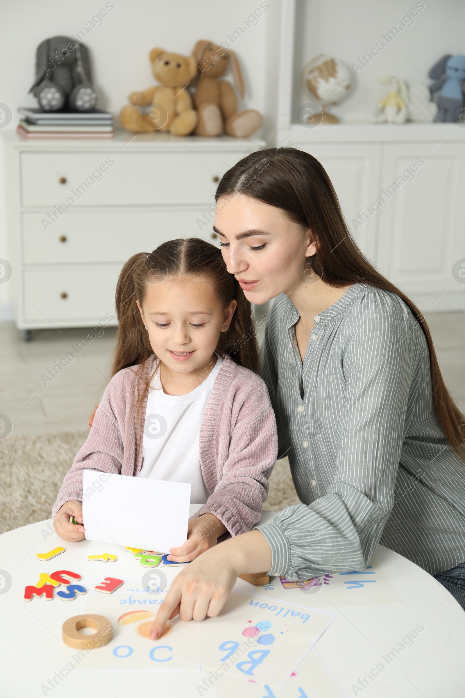 Photo of Speech therapist teaching little girl alphabet at white table indoors