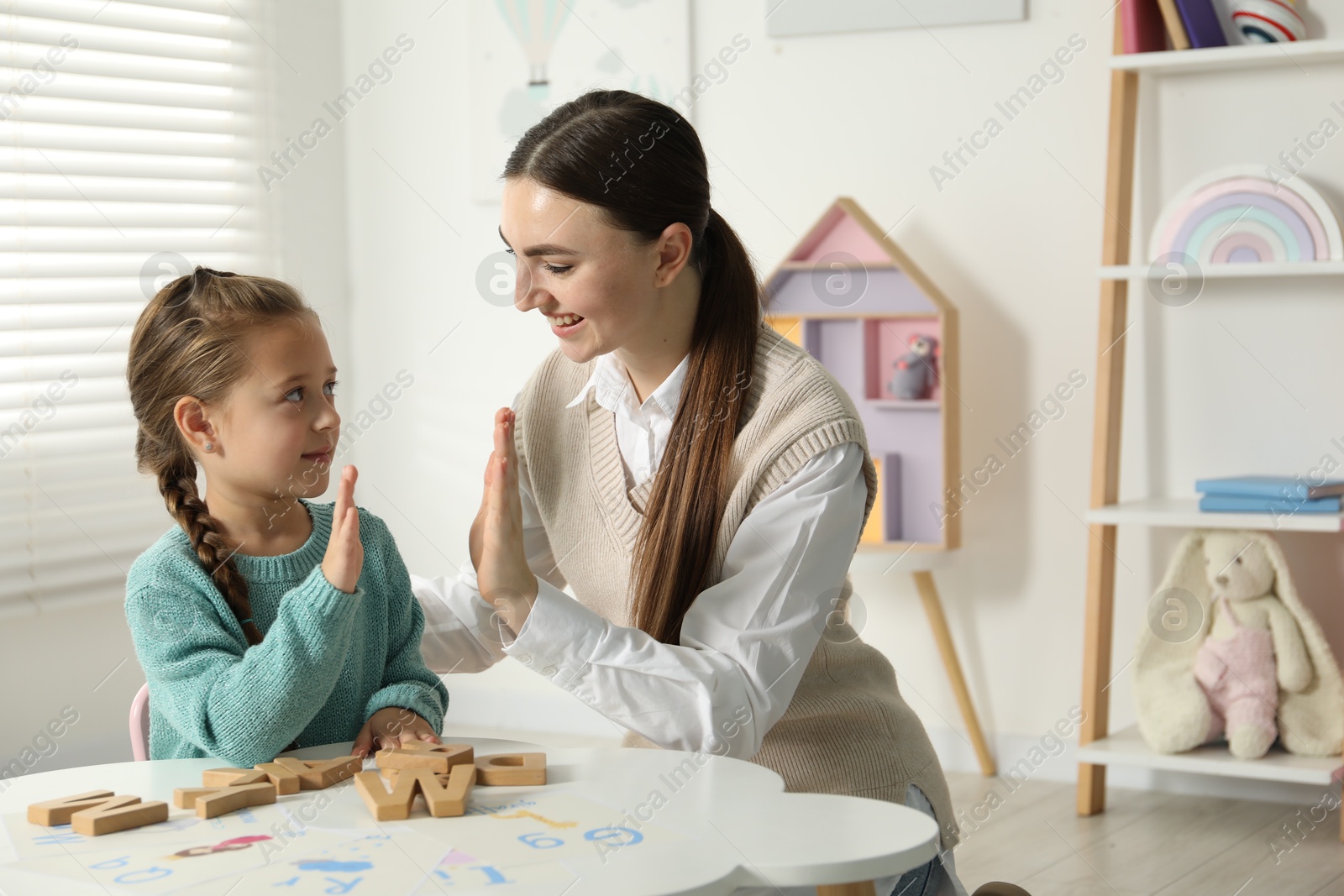 Photo of Speech therapist teaching little girl alphabet at white table indoors