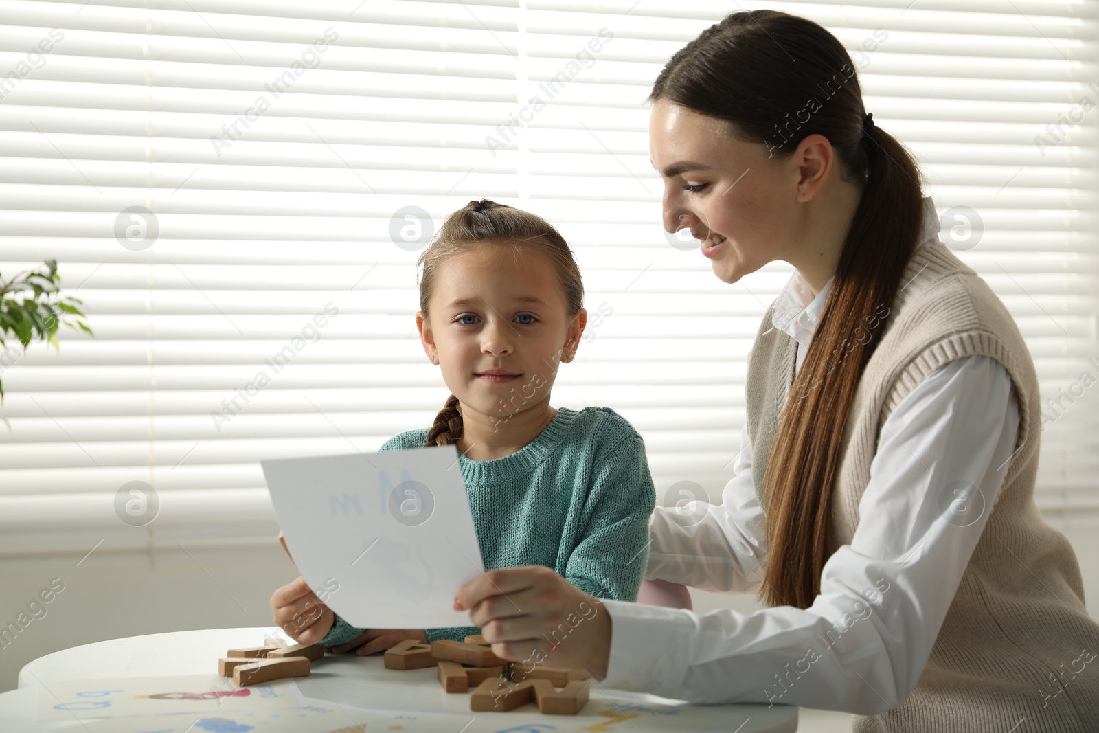 Photo of Speech therapist teaching little girl alphabet at white table indoors