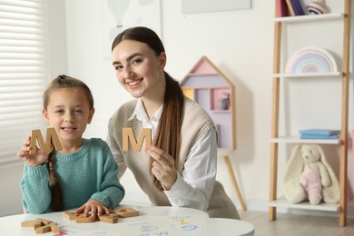 Speech therapist teaching little girl alphabet with wooden letters at white table indoors