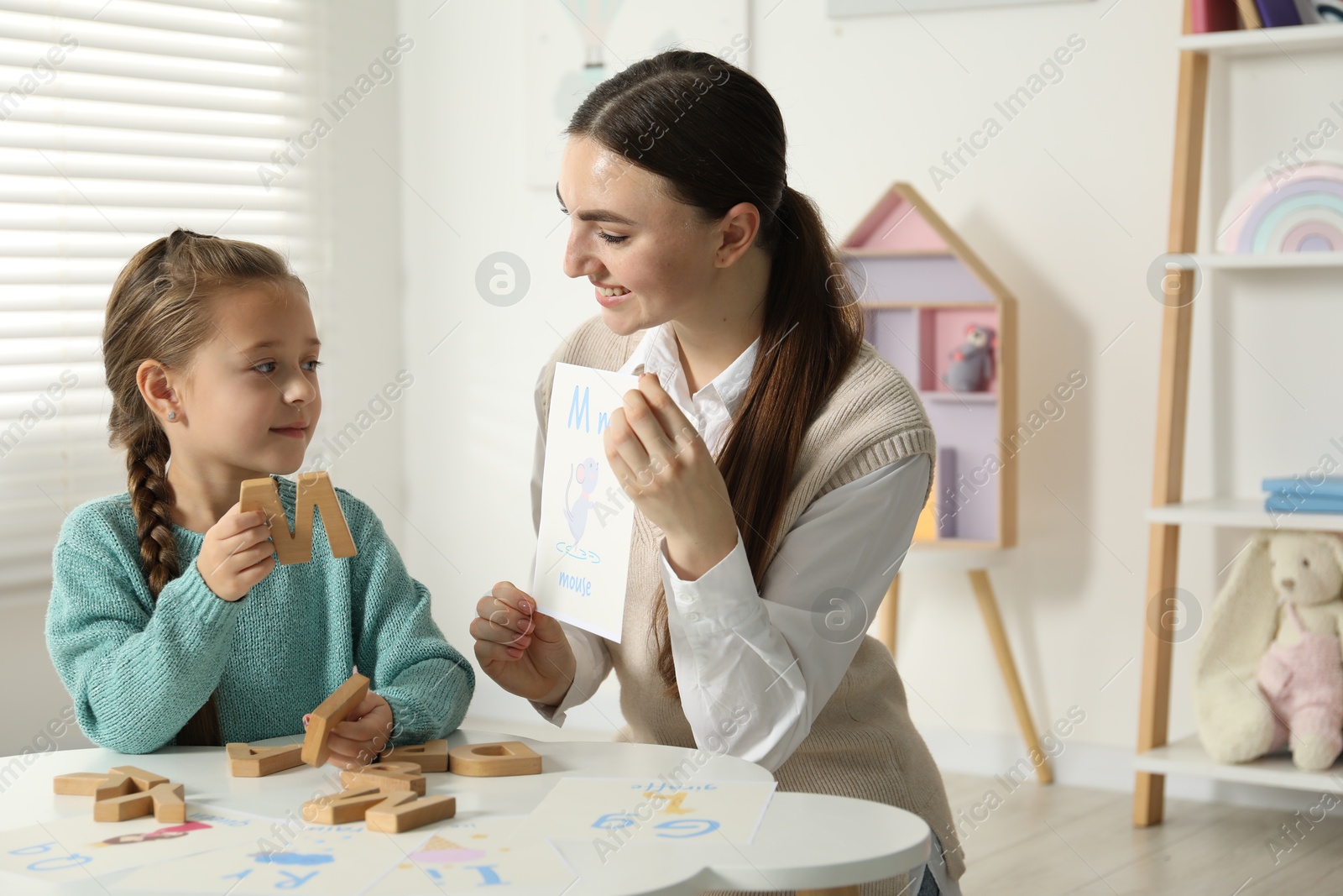 Photo of Speech therapist teaching little girl alphabet at white table indoors