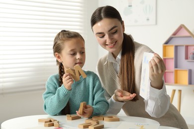 Photo of Speech therapist teaching little girl alphabet at white table indoors