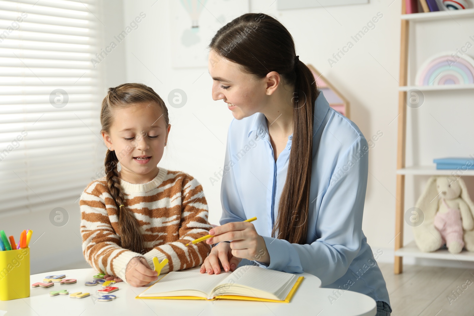 Photo of Speech therapist teaching little girl alphabet with magnetic letters at white table indoors