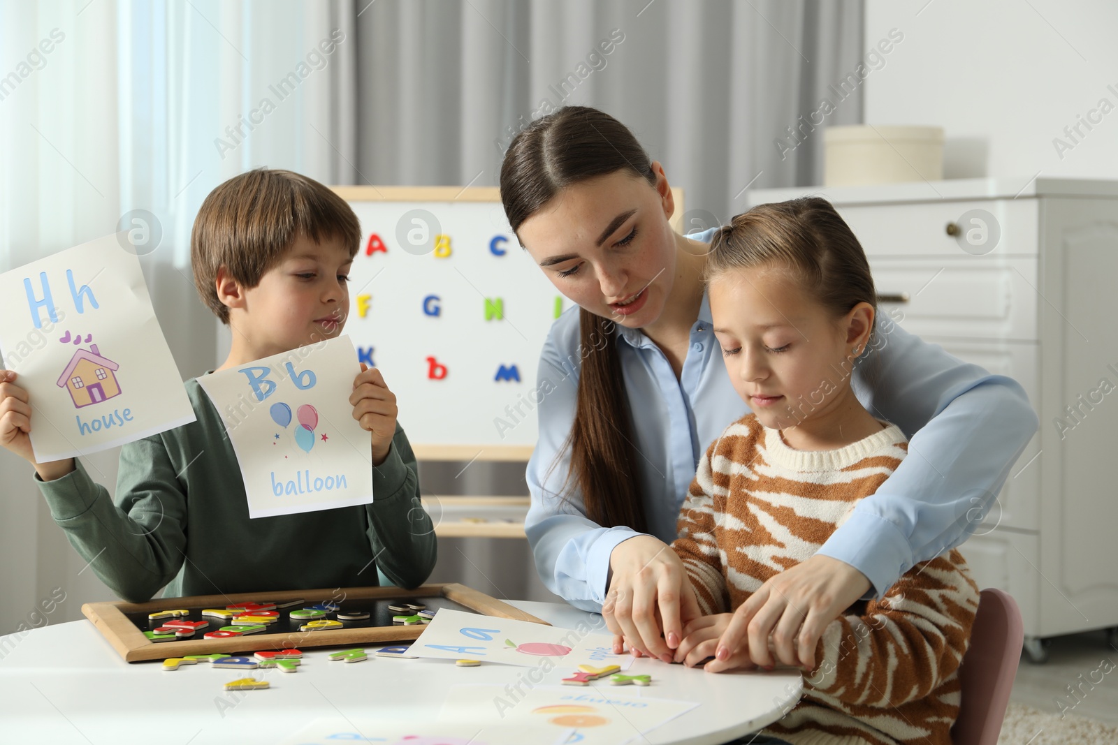 Photo of Speech therapist teaching little kids alphabet with magnetic letters and pictures at white table indoors