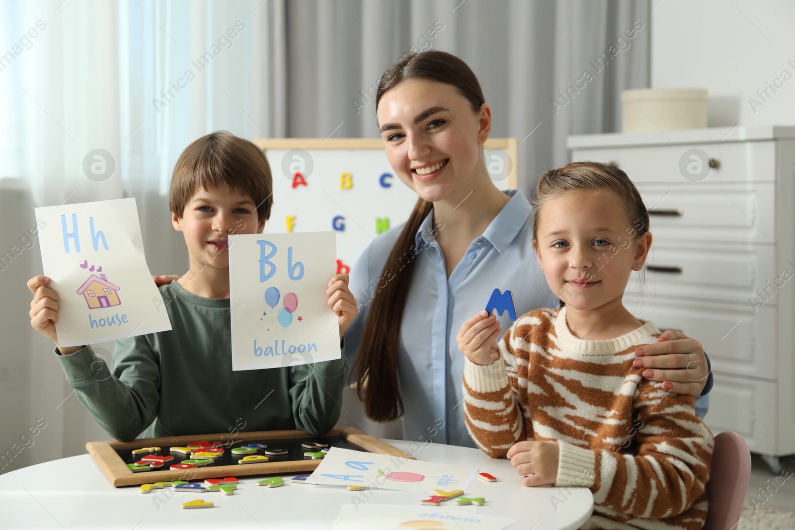 Photo of Speech therapist teaching little kids alphabet with magnetic letters and pictures at white table indoors