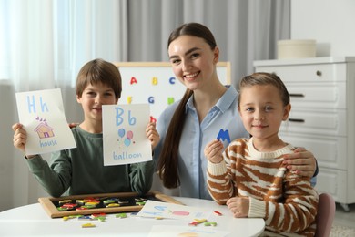 Photo of Speech therapist teaching little kids alphabet with magnetic letters and pictures at white table indoors