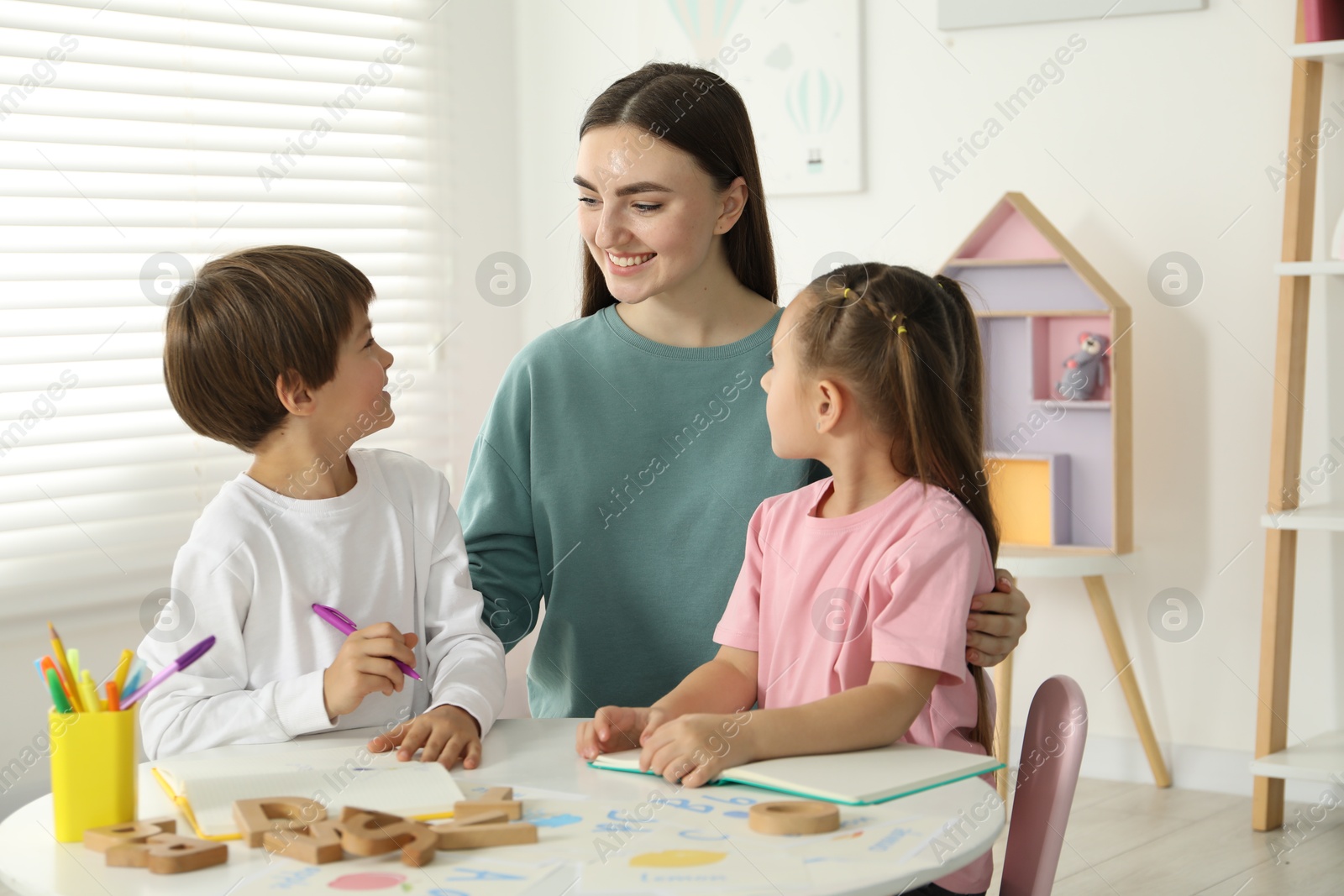 Photo of Speech therapist teaching little kids alphabet at white table indoors