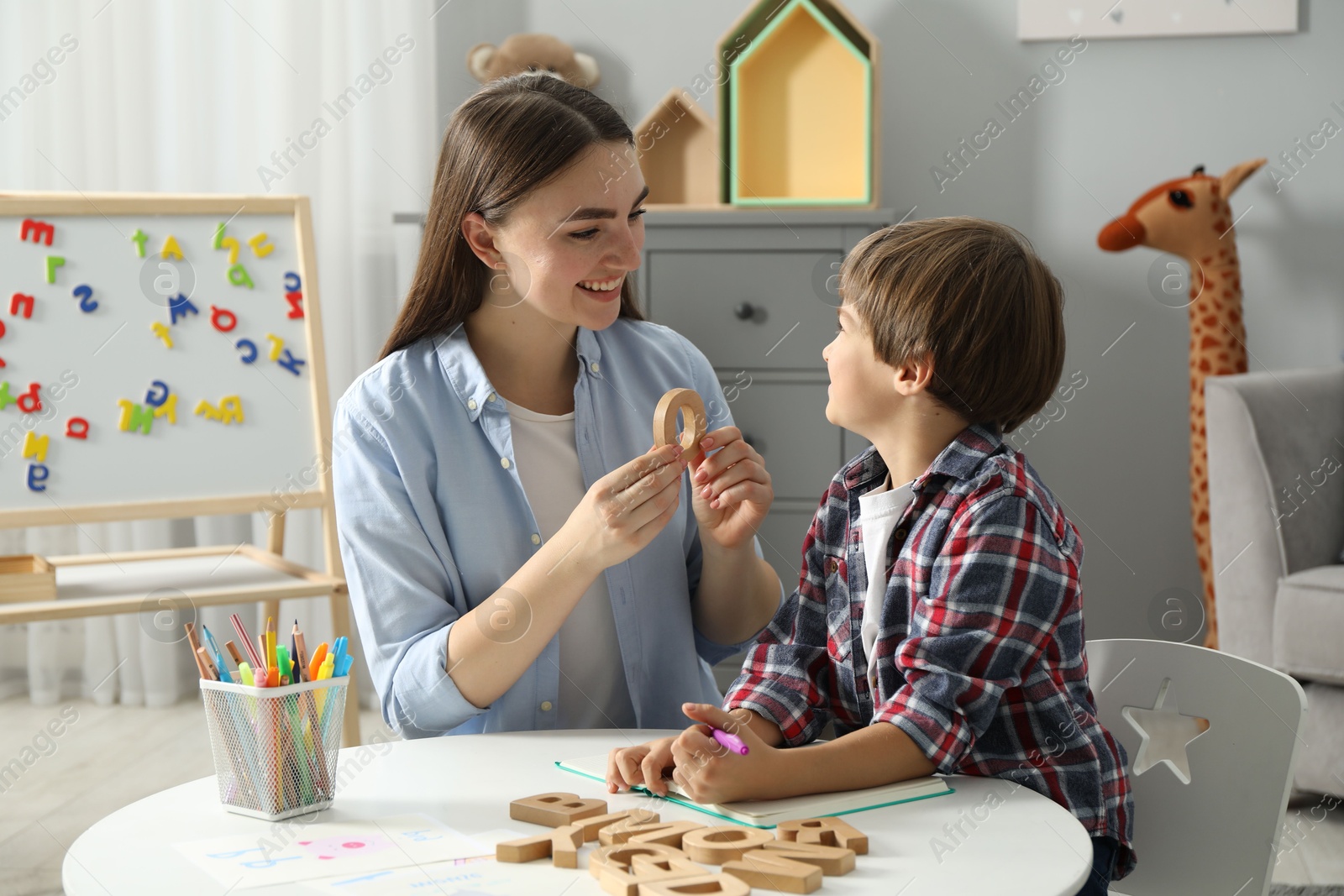 Photo of Speech therapist teaching little boy alphabet with wooden letters at white table indoors