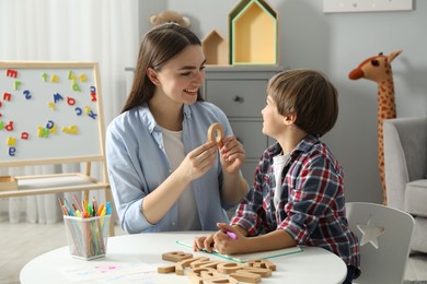 Photo of Speech therapist teaching little boy alphabet with wooden letters at white table indoors