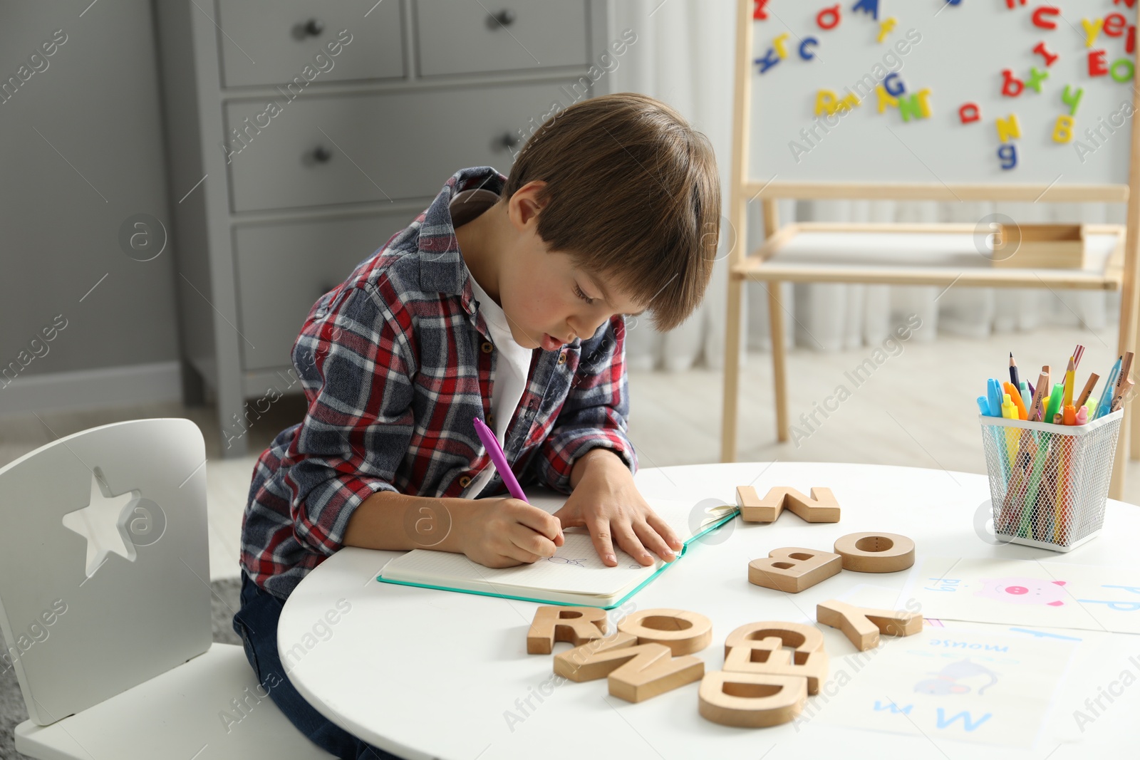 Photo of Little boy learning alphabet at white table indoors