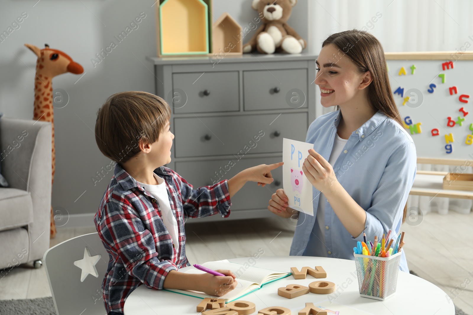 Photo of Speech therapist teaching little boy alphabet at white table indoors