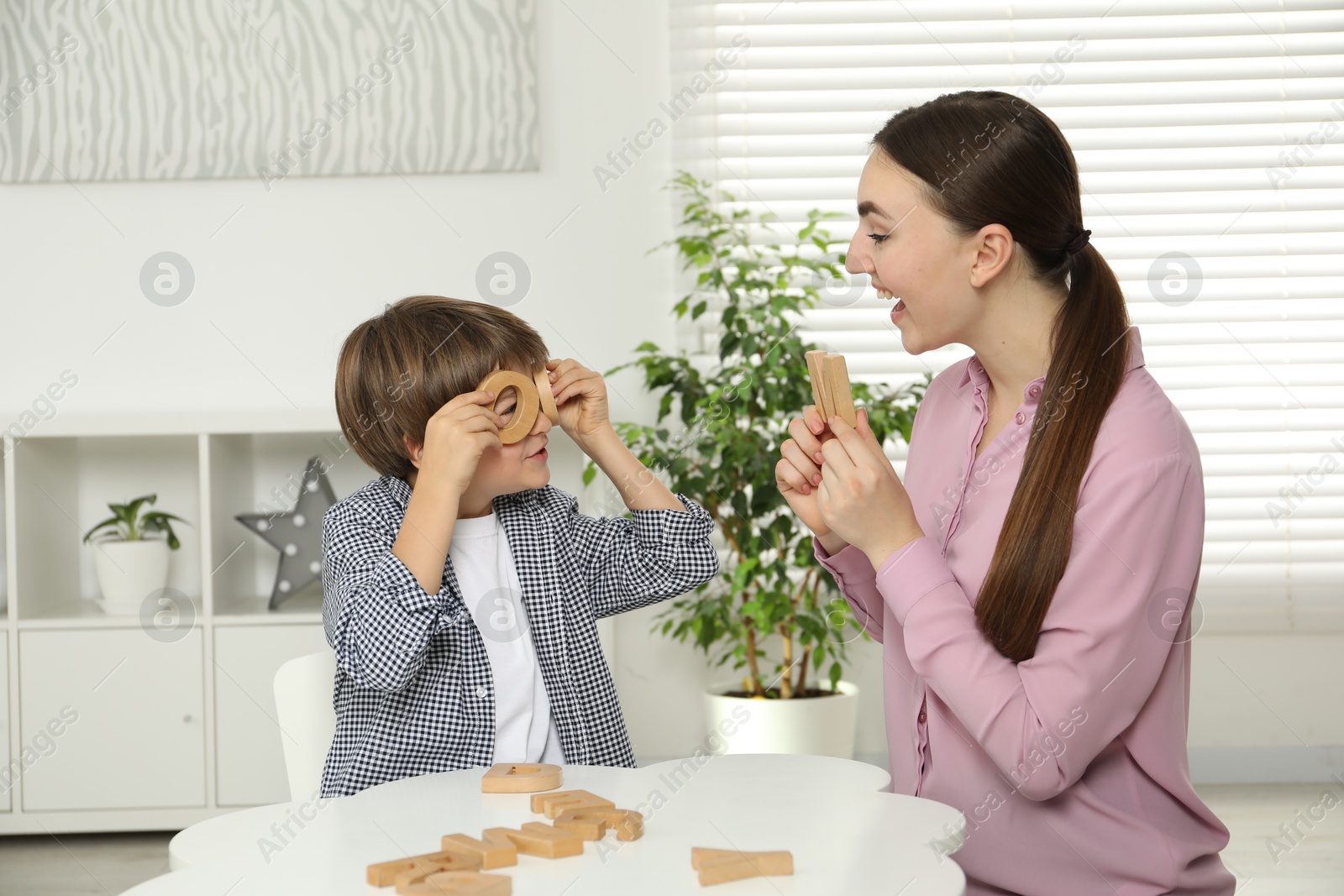 Photo of Speech therapist teaching little boy alphabet with wooden letters at white table indoors