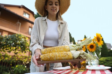 Photo of Beautiful woman with fresh corncobs near table in garden, selective focus