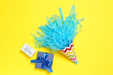 Photo of Party hat with beautiful tissue paper and gift boxes on yellow background, flat lay