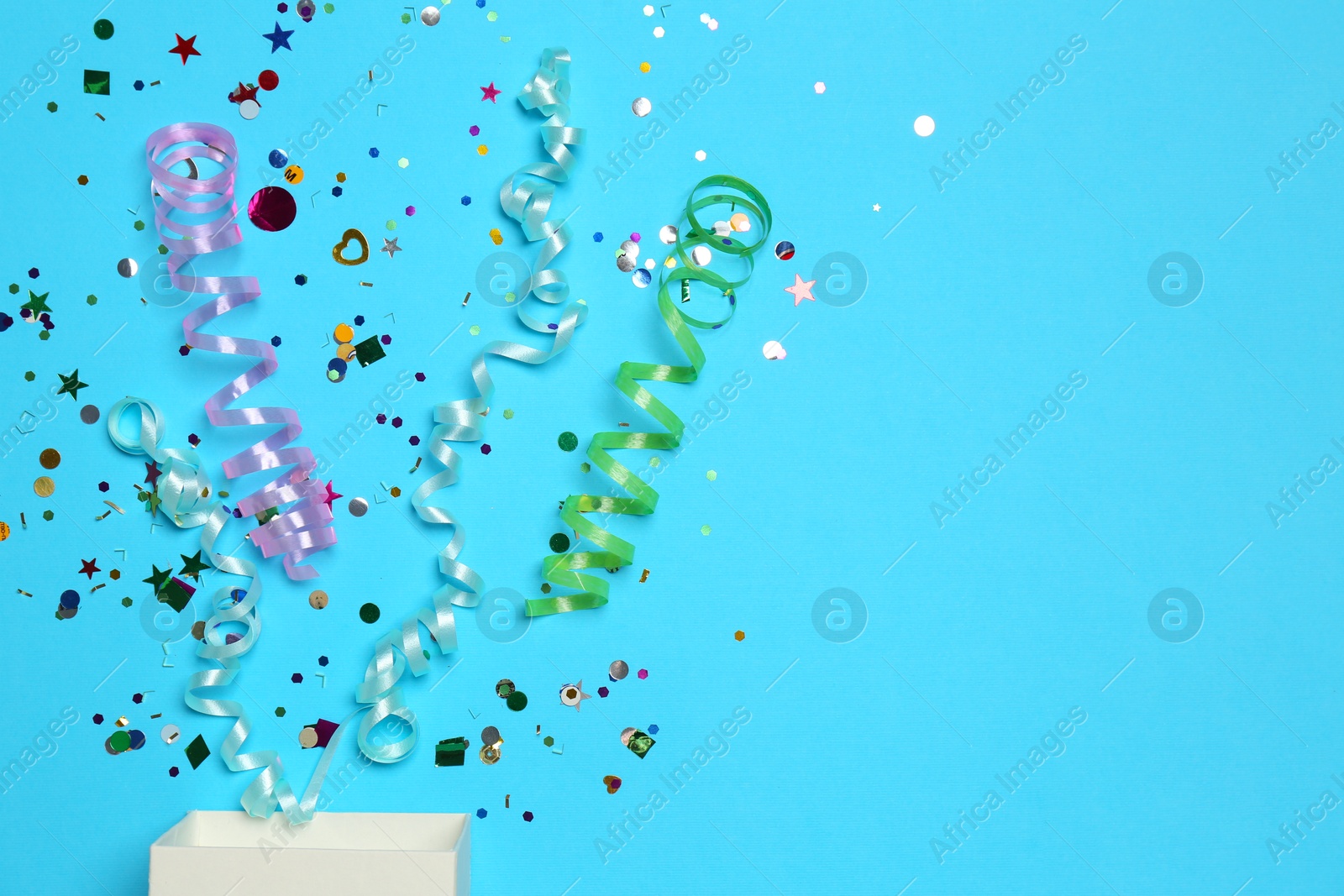 Photo of Shiny confetti and party streamers flying out of little box on light blue background, flat lay. Space for text