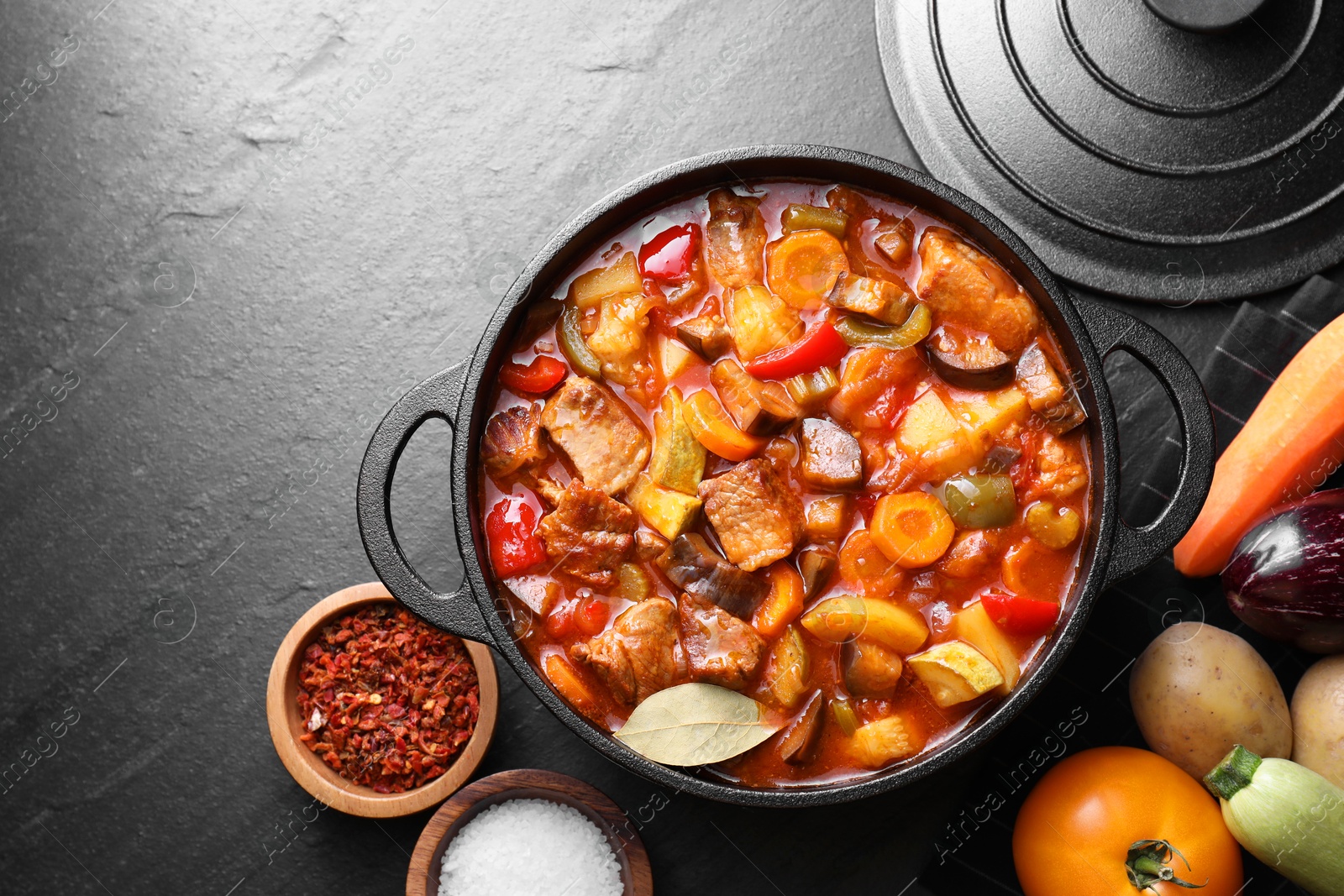 Photo of Delicious stew with vegetables in pot and ingredients on gray textured table, flat lay. Space for text