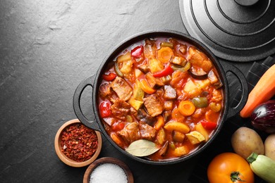 Photo of Delicious stew with vegetables in pot and ingredients on gray textured table, flat lay. Space for text