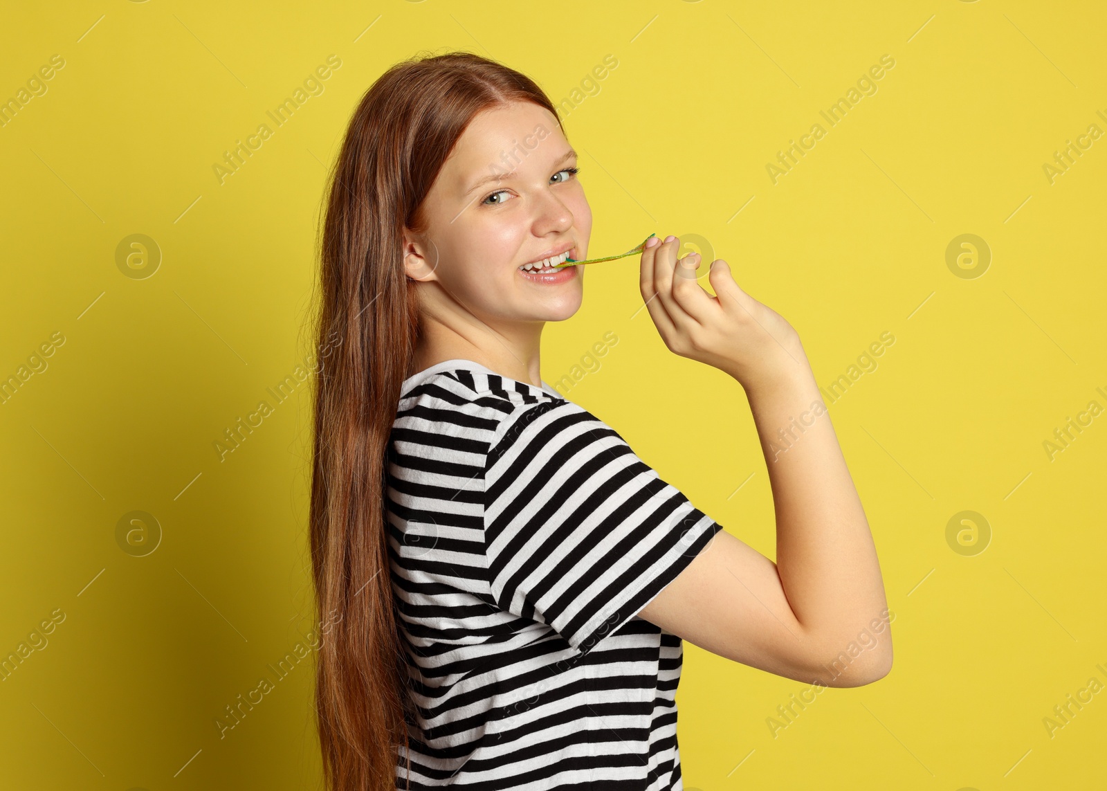 Photo of Teenage girl eating tasty rainbow sour belt on yellow background