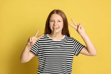 Photo of Teenage girl eating tasty rainbow sour belt while showing v-sign on yellow background