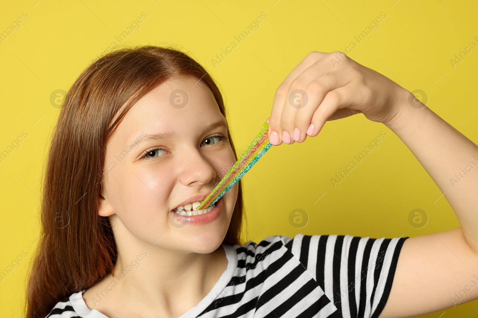 Photo of Teenage girl eating tasty rainbow sour belt on yellow background