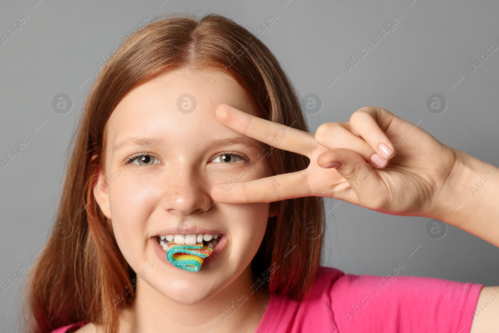 Photo of Teenage girl eating tasty rainbow sour belt while showing v-sign on grey background, closeup