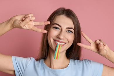 Photo of Young woman with tasty rainbow sour belt showing v-sign on pink background