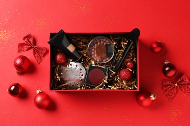 Photo of Christmas gift box with cosmetic products and festive decor on red background, top view