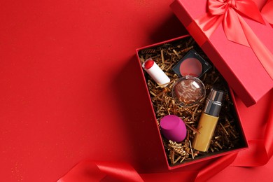 Photo of Christmas gift box with cosmetic products on red background, top view. Space for text