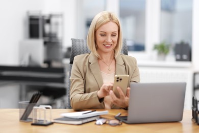 Photo of Smiling middle aged woman using smartphone at table in office